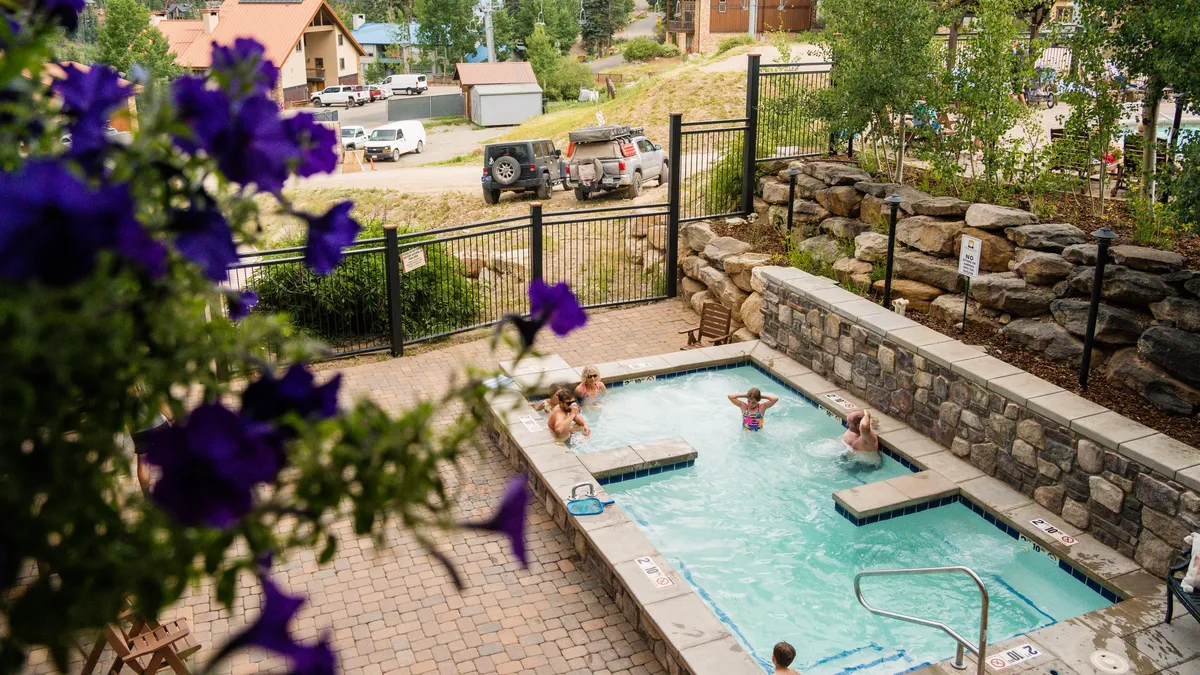 A sparkling outdoor swimming pool surrounded by vibrant flowers, with guests relaxing and enjoying the sunny weather. People are seen swimming and socializing, creating a joyful, inviting atmosphere that suggests relaxation after a day of mountain activities.