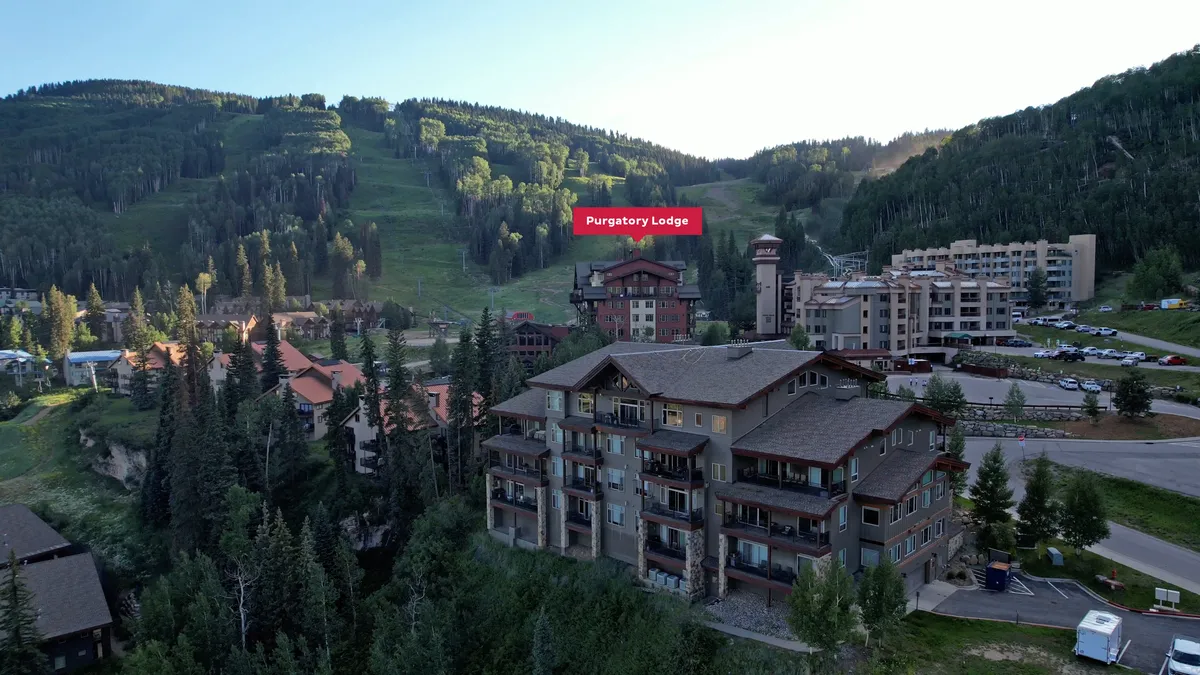 Purgatory Lodge stands prominently in the foreground, framed by lush green mountains and a dense forest area. The image captures the lodge's exterior alongside the expansive natural landscape, with clear daylight inviting viewers to imagine exploring the scenic surroundings. A parking lot hints at easy accessibility and the possibility of embarking on nearby hikes or adventures.