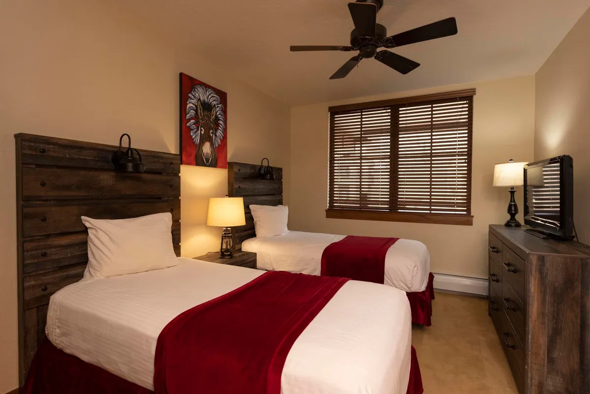 A warm and inviting bedroom with two twin beds adorned with red accents, a lion artwork, a window streaming in morning light, and a TV atop a dresser. The space is set up for both comfort and togetherness, ideal for families or groups.