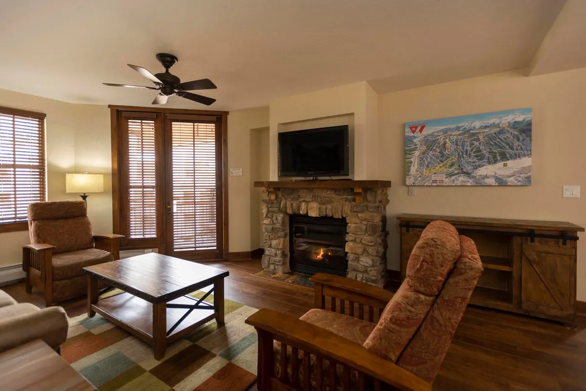 The image showcases a cozy, warmly lit living area featuring a stunning stone fireplace as the centerpiece, surrounded by plush, comfortable chairs and a wooden coffee table. A TV is mounted above the fireplace, making the room perfect for relaxing evenings, family gatherings, or quiet mornings. The ambiance is inviting and exudes mountain lodge luxury.
