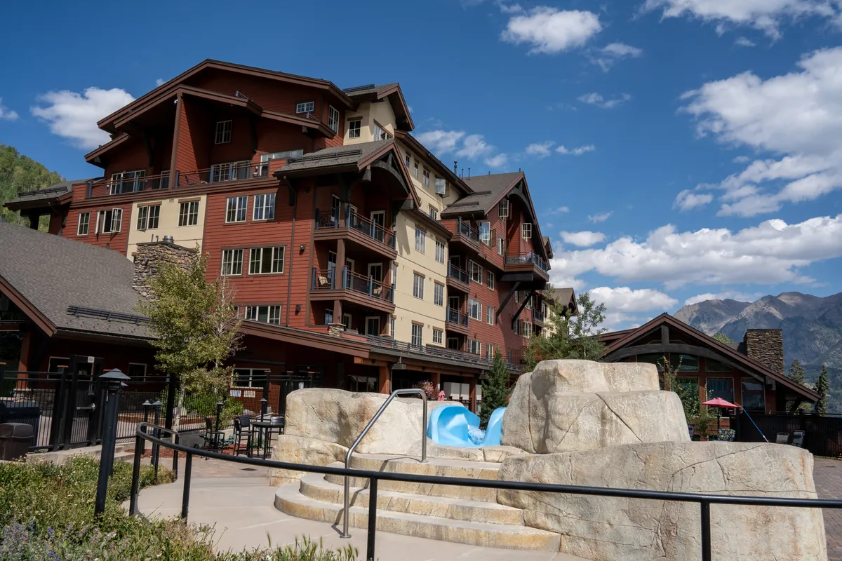 This image captures a lively exterior view of the swimming pool area, complete with a fun water slide, stone structures, and the multi-story lodge building in the background. Guests are swimming, sunbathing, and socializing, creating an atmosphere of leisure and community.