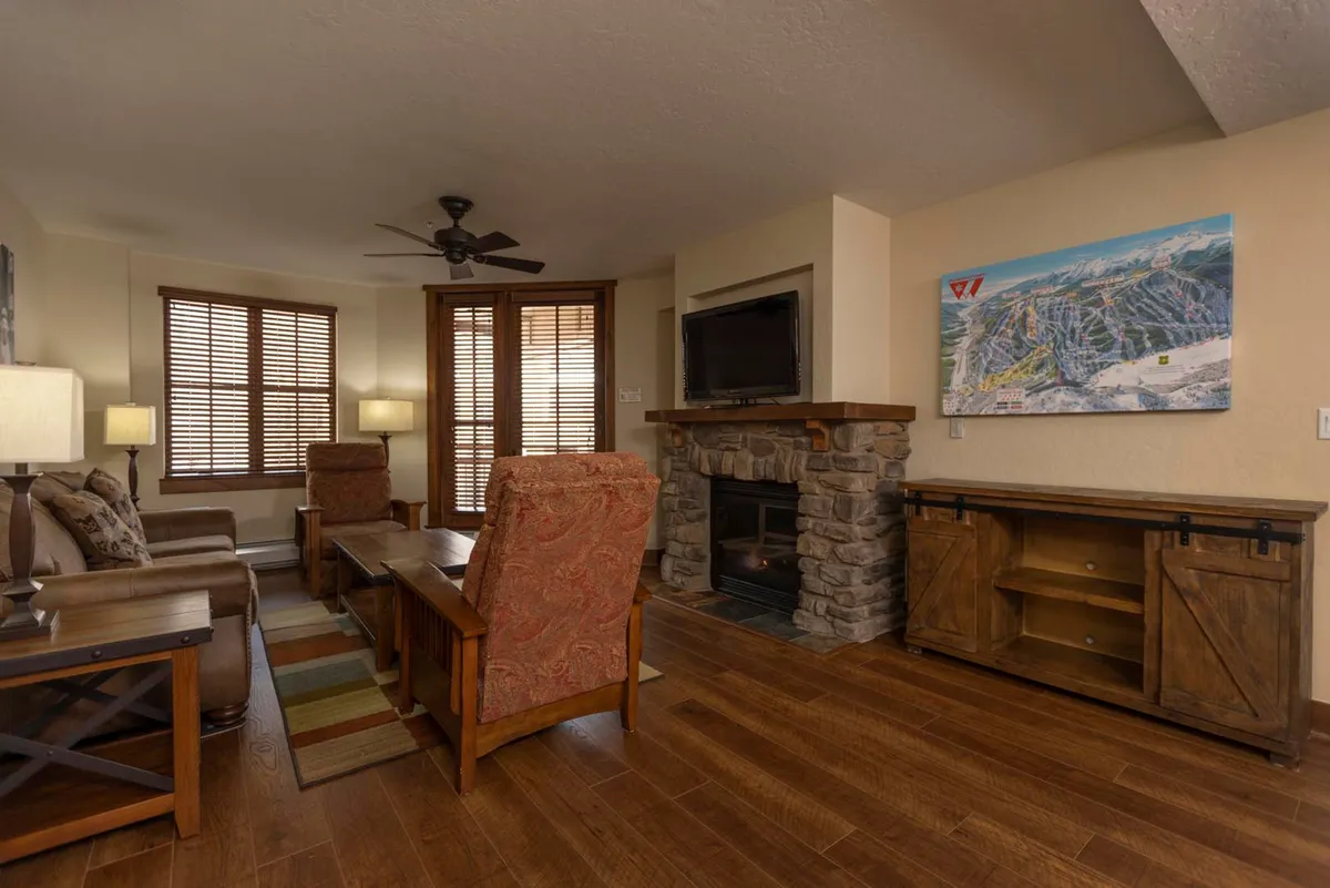 A cozy living room featuring a prominent stone fireplace surrounded by comfortable seating, with artwork on the wall and natural light filtering in through nearby windows. The room exudes warmth and invites relaxation during chilly mountain evenings.