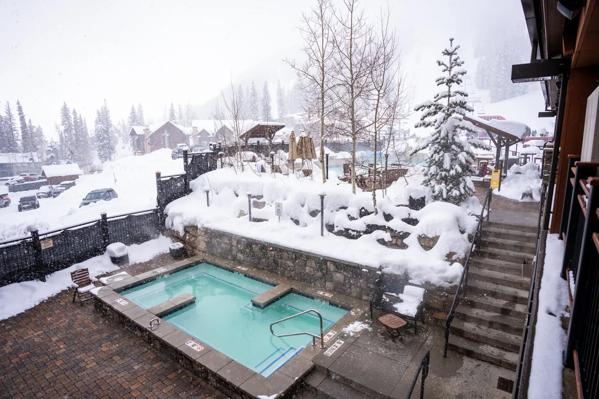 A serene outdoor hot tub is nestled in a snow-covered landscape, surrounded by trees and a staircase. Guests relax in the bubbling water, enjoying a peaceful contrast between the warmth of the spa and the wintry surroundings.