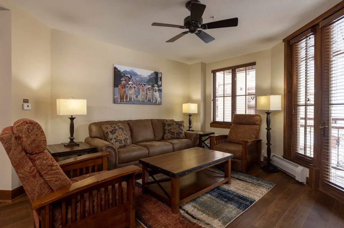 This image features a warm, welcoming living area with a plush sofa, coffee table, elegant artwork, and large windows that likely frame mountain views. The soft lighting from table lamps and the relaxed arrangement create an inviting lounge atmosphere ideal for unwinding after a day of indulgence.