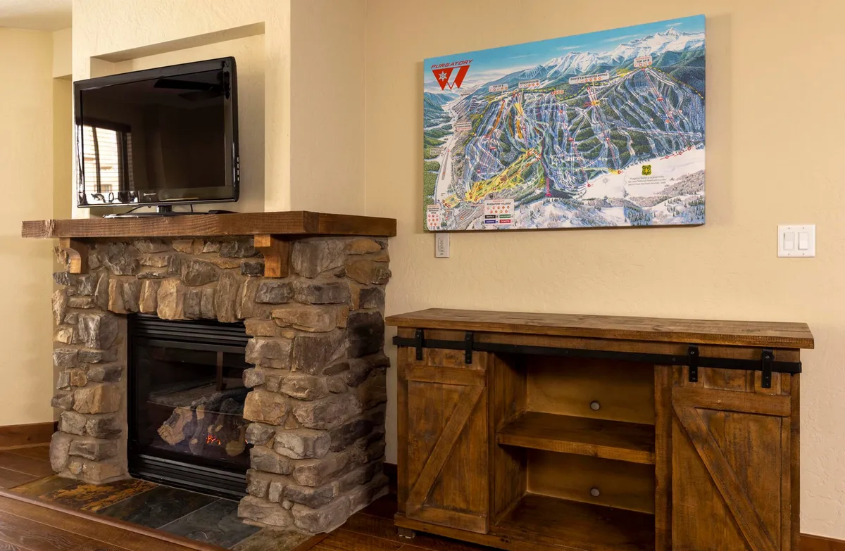 A warm, welcoming interior with a stone fireplace, a wooden console table, TV, and a large ski trail map on the wall. The scene suggests post-ski relaxation, family gatherings, and a sense of connection to the mountain setting.
