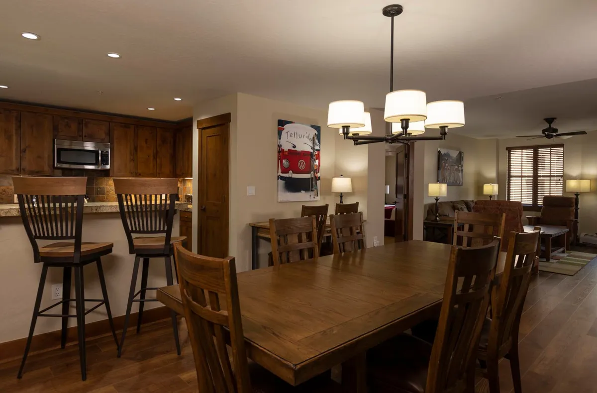 An inviting open-plan dining area featuring a spacious wooden dining table under a chandelier, bar stools at the kitchen island, and visible kitchen appliances. Artwork adds a touch of personality, and the space is set up for family dinners, game nights, and entertaining guests.