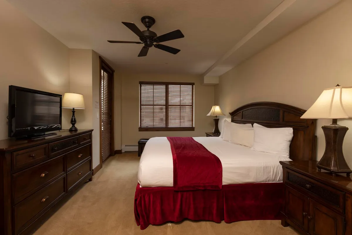 A serene bedroom featuring a plush bed with a red accent blanket, a wooden dresser, and a window with blinds allowing in soft natural light. The room exudes relaxation with its cozy setup, ideal for unwinding after a day in the mountains.
