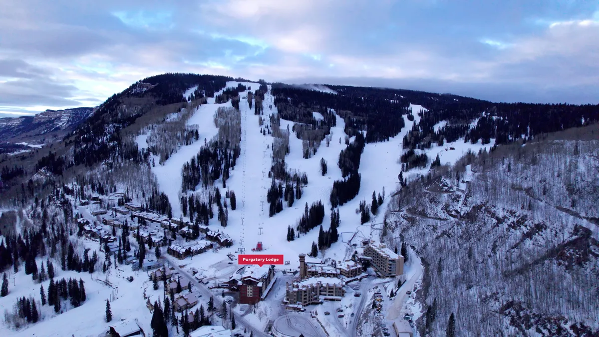 A panoramic view of Purgatory Lodge nestled at the base of snow-covered ski slopes, framed by majestic mountains and frosted trees. The scene radiates the excitement of a winter day with fresh powder, with skiers gliding down the slopes and the inviting lodge at the heart of the action.