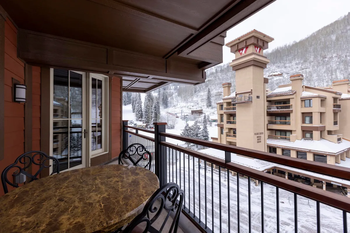 The image shows an outdoor dining table set against a stunning mountain backdrop, with snow gently falling. People are gathered around the table, enjoying breakfast or sipping hot cocoa, while an adjacent building hints at the proximity to the lodge and amenities. The mood is inviting and warm, highlighting the joy of being together in a scenic mountain setting.
