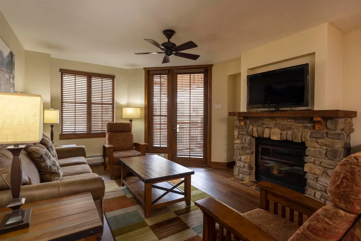 A warm, welcoming living room with a prominent stone fireplace, wooden furniture, a TV, and a large window with blinds letting in natural light. The space invites relaxation and socializing, with cozy touches like a decorative lamp and a plush seating arrangement.