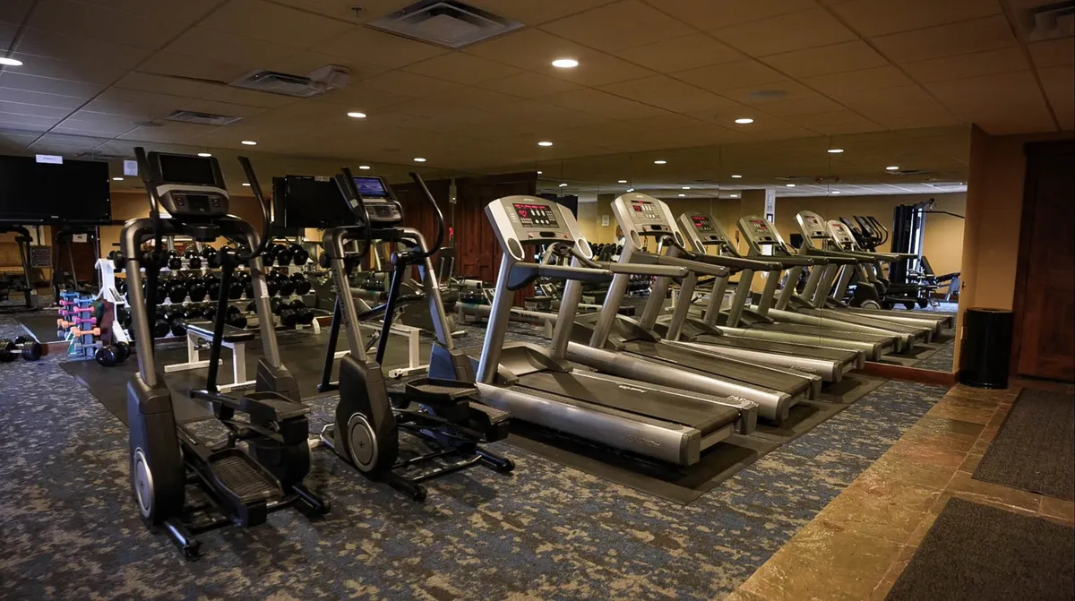 Inside the state-of-the-art fitness center, treadmills, ellipticals, dumbbells, and mirrors provide a modern, energizing environment for maintaining wellness routines. The room is spacious and filled with natural light.
