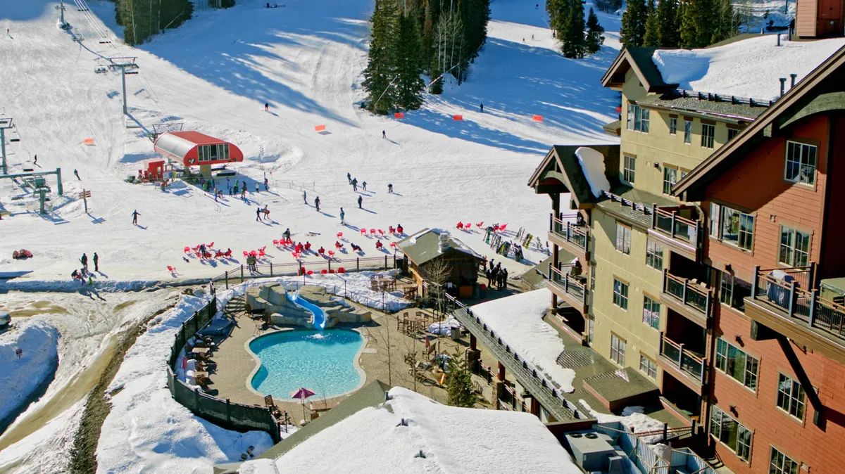 The scene captures the lively exterior of the lodge, with skiers and snowboarders on the slopes and people swimming and relaxing by the pool. The lodge itself is visible, anchoring the activity and providing a sense of place. The mood is energetic and festive, showcasing both winter sports and leisurely swims within close proximity.